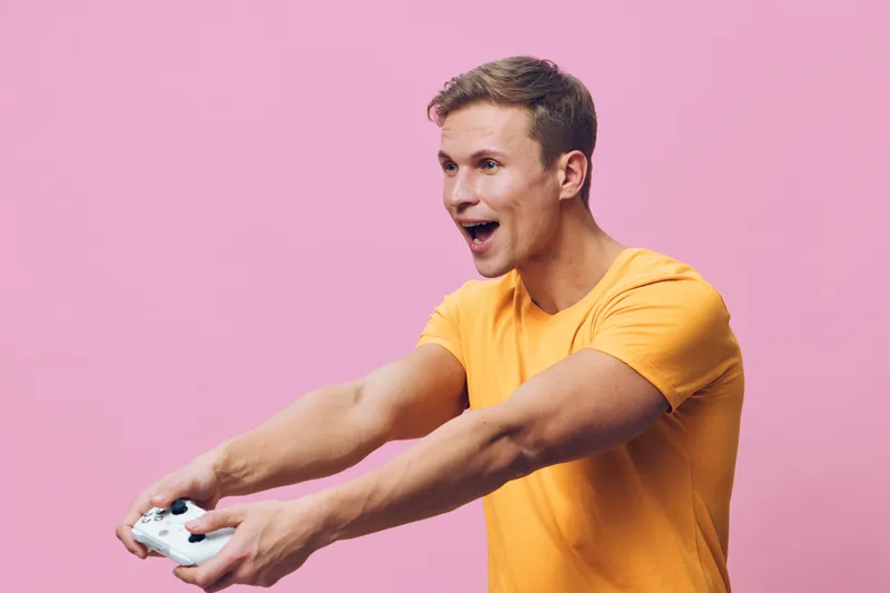 Energetic young man with short brown hair wearing orange t-shirt playing video game holding gamepad with excitement against pink background studio portrait. People entertainment concept