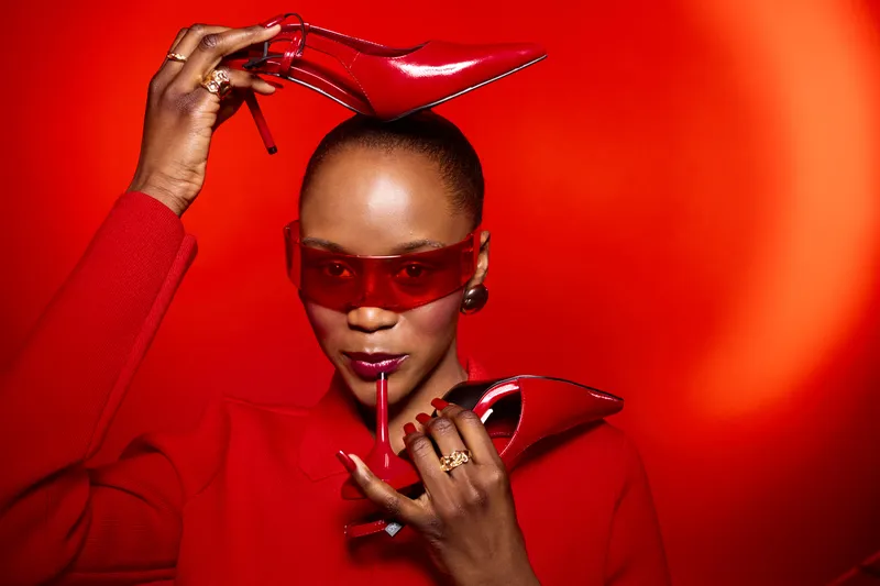 Stylish dark skinned African woman in red sweater and sunglasses poses with red high heels on red background. Concept of fashion, beauty, elegance, and bold color styling.