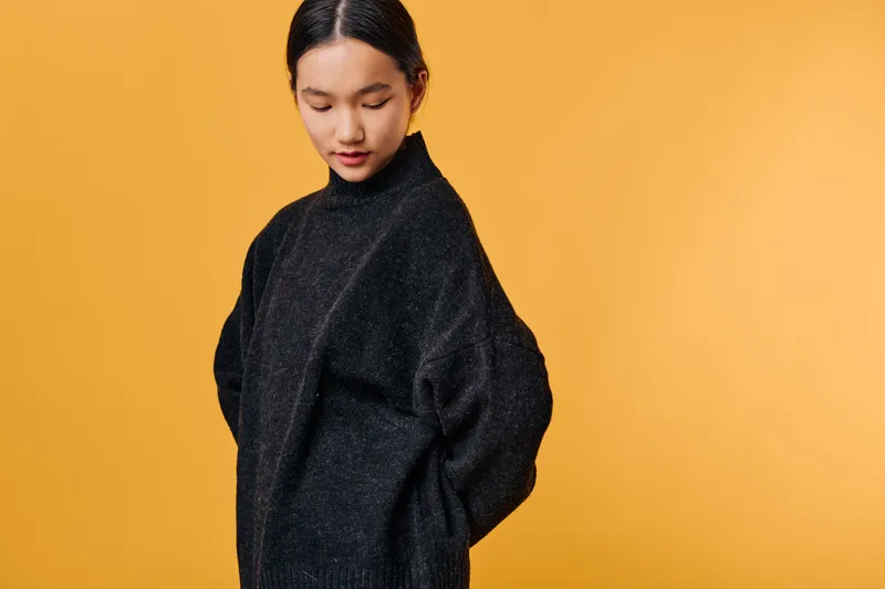 Young woman with dark hair wearing oversized black sweater standing against orange background looking down thoughtfully with hands behind back in casual fashion portrait.