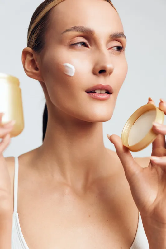 Young woman applying cream on her cheek, bright studio lighting, minimal beauty shot, skincare routine moment, soft moisturizer texture, natural tones and calm expression