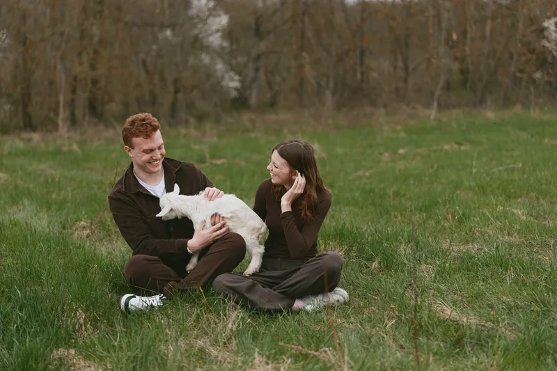 Smiling couple with a goat kid in a grassy field, enjoying outdoor time together. Warm natural light highlights companionship, playfulness, and gentle animal interaction in a peaceful countryside