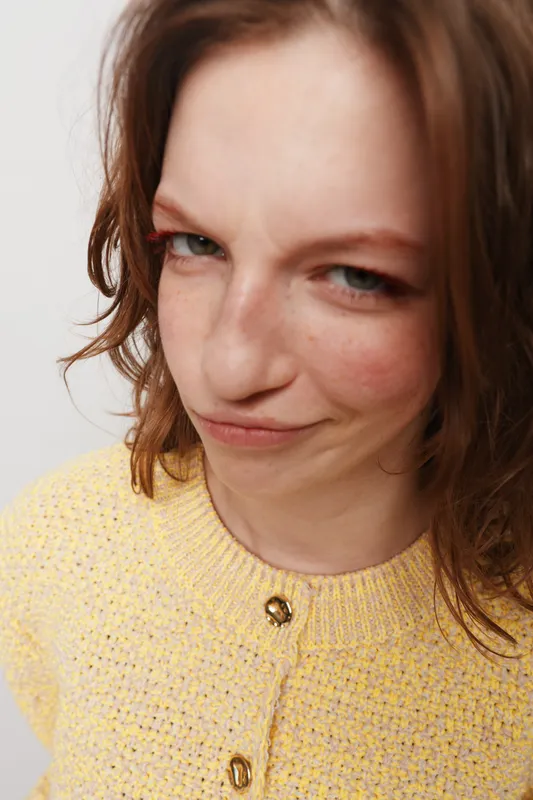 Close-up of a young woman with brown hair wearing a yellow knitted sweater making a playful smirk. The white background highlights her natural expression and casual style.