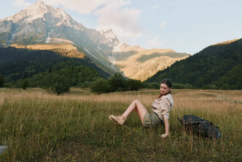 Woman traveler relaxing in a meadow with distant mountains, wearing hiking clothes, sunlit outdoor scene, capturing a calm nature lifestyle moment in landscape