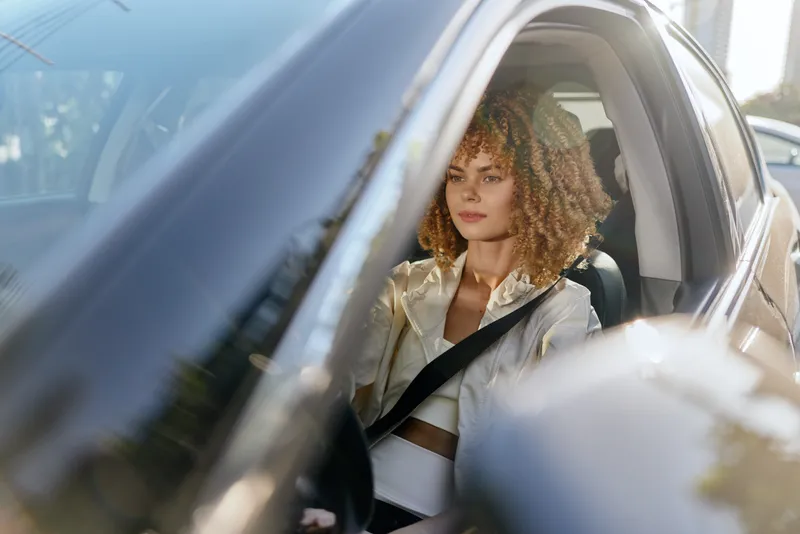 young woman driving car with seatbelt fastened, focused on road during daytime, curly hair, casual outfit, city background, natural light