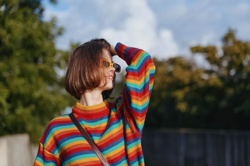 Woman in a colorful striped sweater with sunglasses and short hair, standing outdoor in a city park, smiling and shielding her eyes with hand, casual outfit and lifestyle fashion.