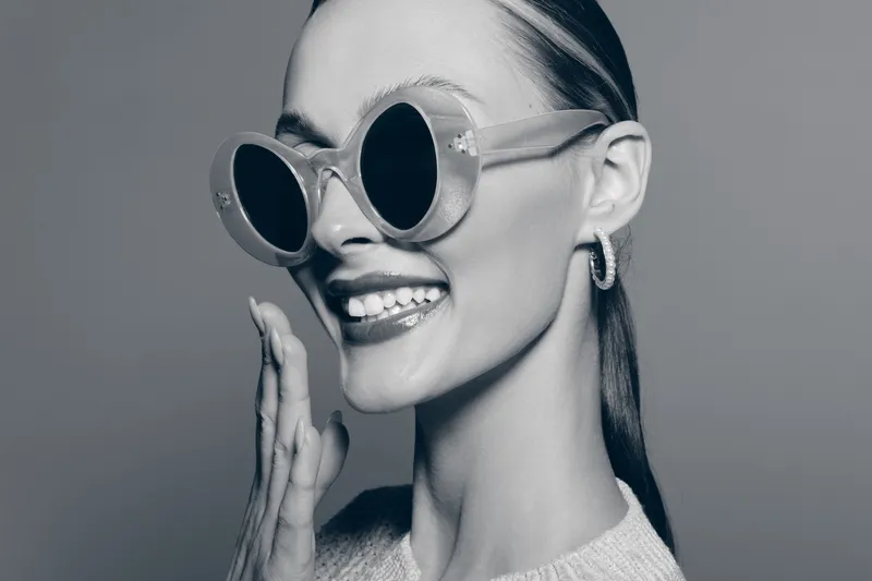 Smiling white woman with fair skin wearing round sunglasses and hoop earrings in studio. Fashion and beauty concept.