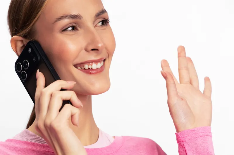 Smiling young woman in pink sweater talking on smartphone, waving with hand. Bright studio background, natural makeup, showing happiness and communication concept.