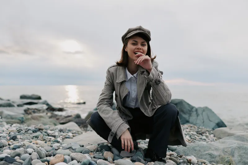Young woman wearing a gray coat and cap squats on a rocky beach near the water during cloudy weather. Casual style, outdoor, fashion, nature, and coastal concept.