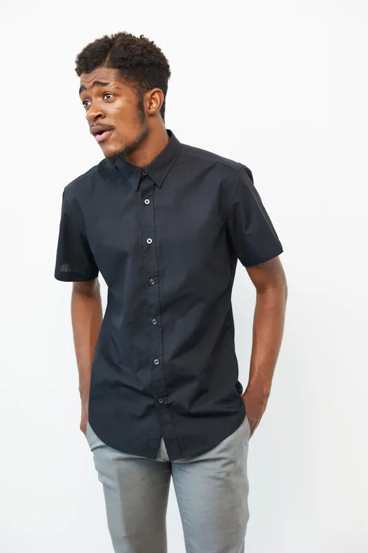 Stylish black man wearing a black shirt and gray pants, posing with a confident expression against a minimalistic white background.