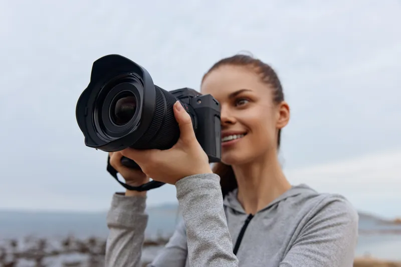 Young woman photographing outdoors, showcasing passion for photography with a DSLR camera, wearing a grey hoodie in a serene coastal setting