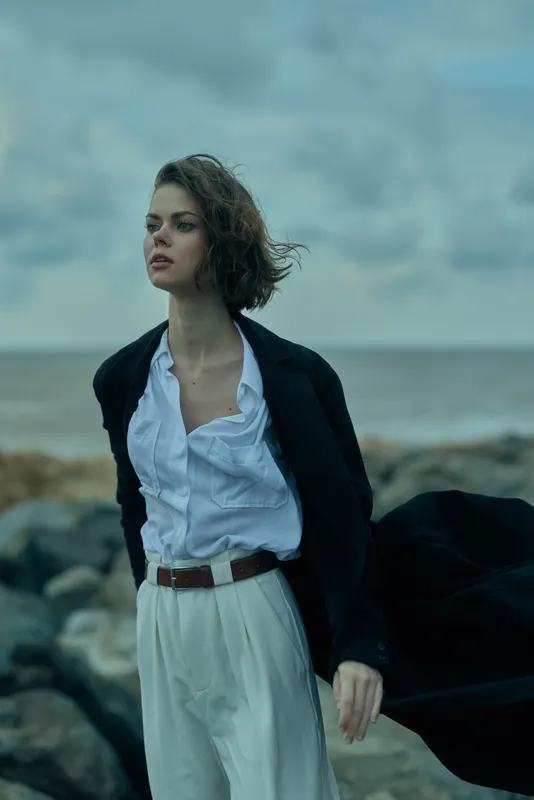 Woman stands on a windy coast wearing a blazer over a white shirt and belted white trousers, gazing into the distance as waves crash behind.