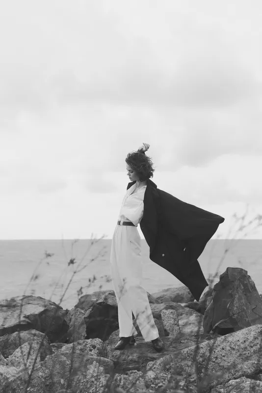 Woman stands on rocky coast with cape billowing, sea in background, monochrome mood, fashion statement, sense of freedom and bold movement
