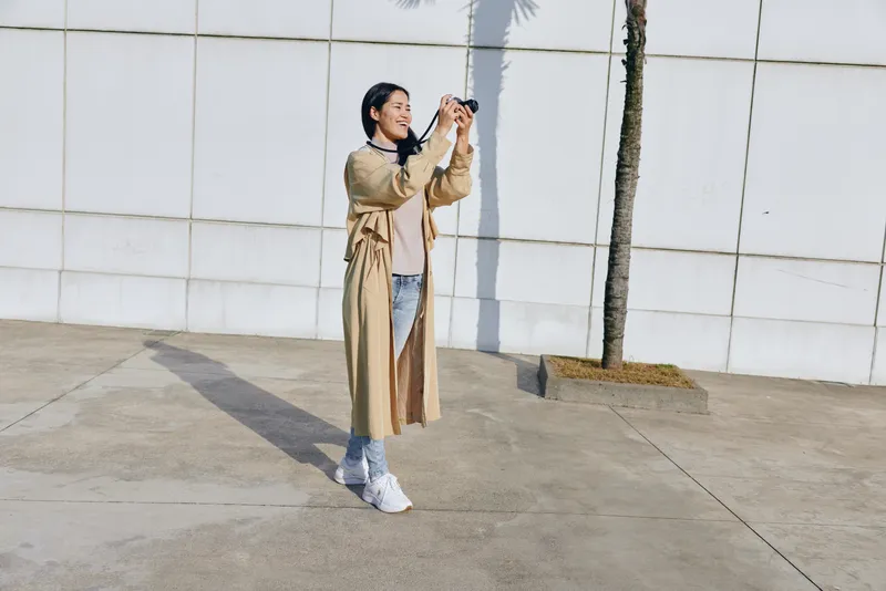 Photography of a woman in a beige trench coat outdoors, capturing a moment with a camera, urban backdrop, casual street fashion, sunny day, candid mood.