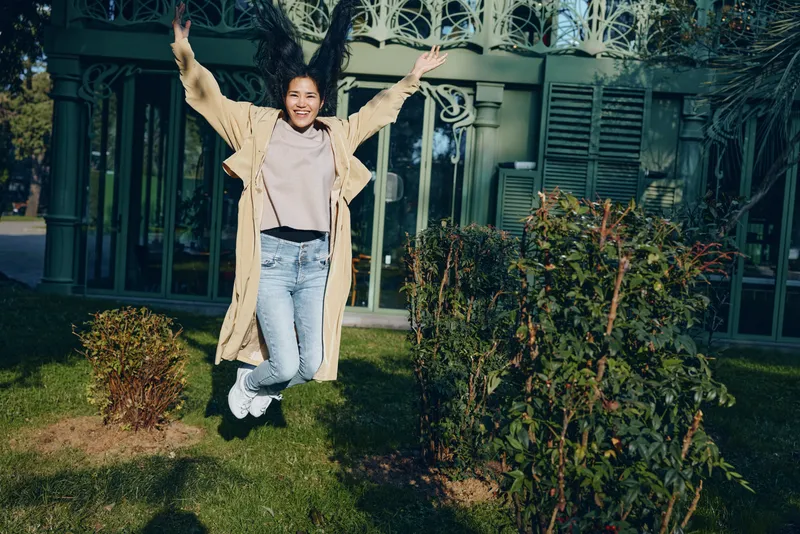 Joyful outdoor jump in a green garden with bushes and a house in background, runway casual coat, jeans, sneakers, carefree season moment captured in daylight, energetic movement and happiness during