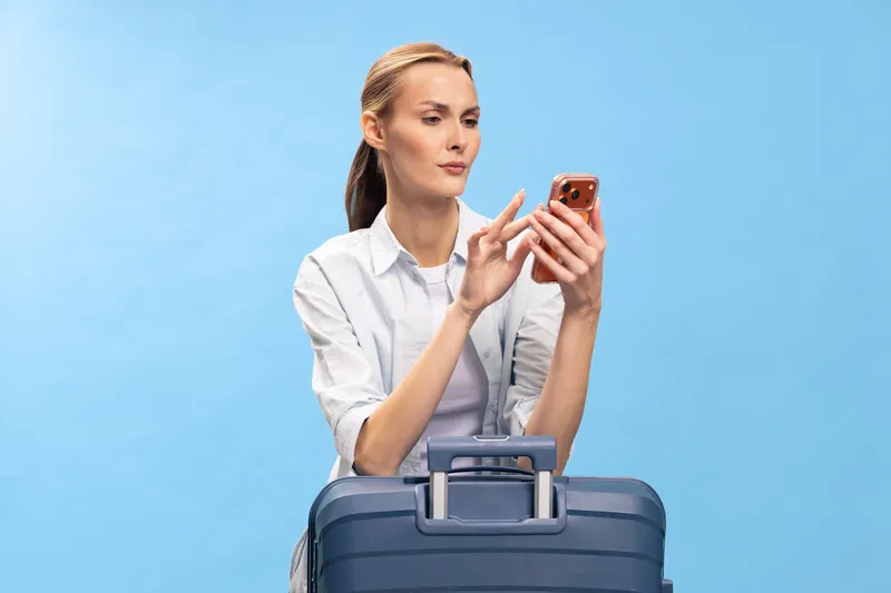 Young woman in white shirt holding smartphone stands behind blue suitcase on blue background. Concept of travel planning, vacation preparation, and modern communication technology.