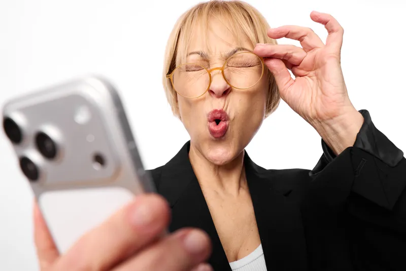 Woman adjusts glasses with one hand while holding a smartphone, blonde businesswoman looks surprised, office background, blurred phone foreground, indoor portrait.