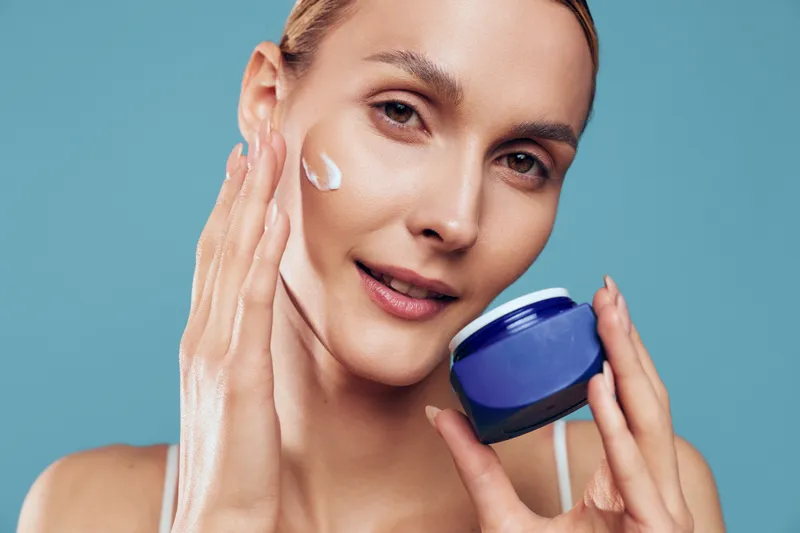 Woman applying face cream on cheek with hand holding blue jar. Close-up portrait on blue background. Concept of skincare, beauty routine, and moisturizing skin care.