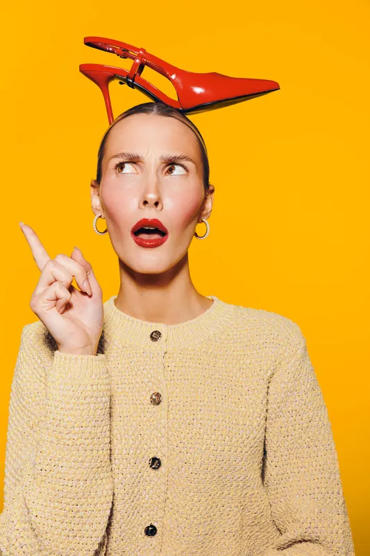 Young woman with red high heel shoe on head looks surprised and points up. Bright yellow background and beige sweater create contrast. Concept of fashion, creativity, and expression.