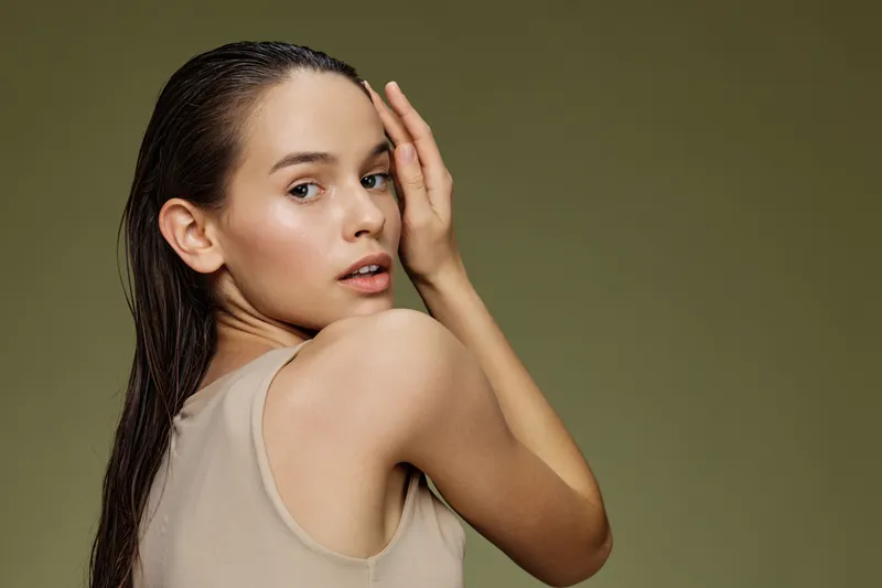 Young woman with wet hair looking over her shoulder, wearing a neutral tank top against a green background, conveying a fresh and natural beauty concept