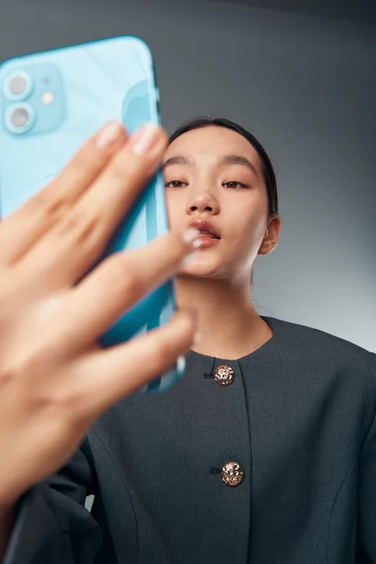 Young woman taking selfie photo with smartphone in modern outfit, closeup portrait with focused expression and casual background for social media use.