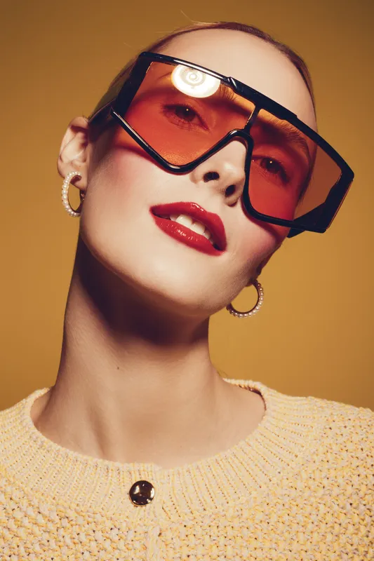 Close-up portrait of a stylish woman wearing oversized orange sunglasses and hoop earrings. She has red lipstick and a textured beige sweater on a warm yellow background. Fashion concept.
