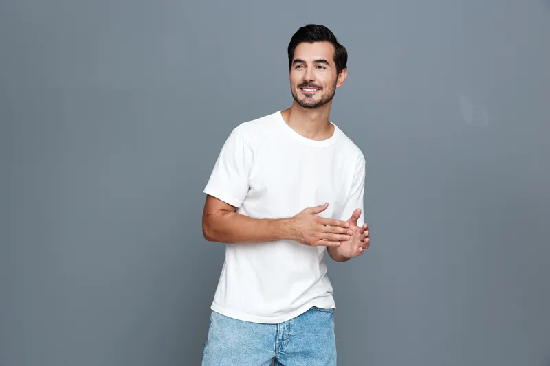 Happy cheerful young man wearing a white t-shirt and blue jeans stands on a solid gray background, smiling and gesturing with his hands in a friendly manner.