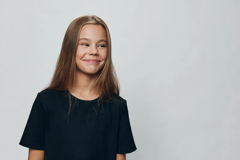 Girl with long hair wearing black t shirt is standing and smiling on light neutral background looking sideways with playful and happy expression in studio portrait.