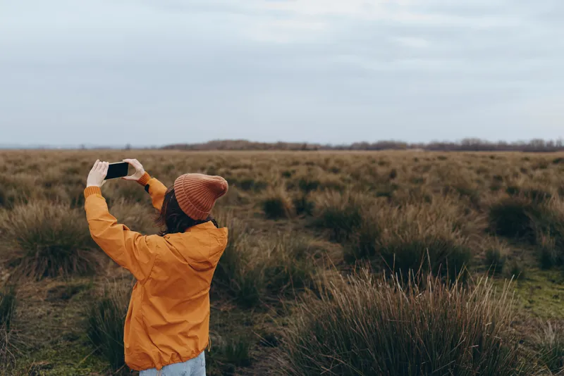 Woman outdoors lifestyle in a grassy field landscape, orange jacket, arms raised, facing the horizon, nature calm wind day, standing alone in countryside, travel photo portrait mood