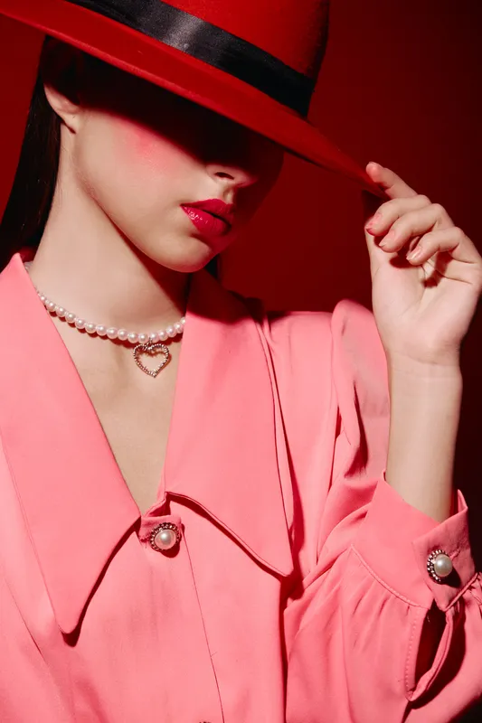 Portrait of stylish woman in a pink blazer and red hat partially covering face. Makeup includes glossy lips, with soft light highlighting elegant pearl necklace. Fashion and beauty concept.