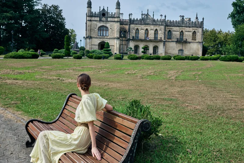 woman on bench in park facing historic mansion, authenticity inclusivity, long vintage dress, contemplative pose, landscaped lawn, overcast sky