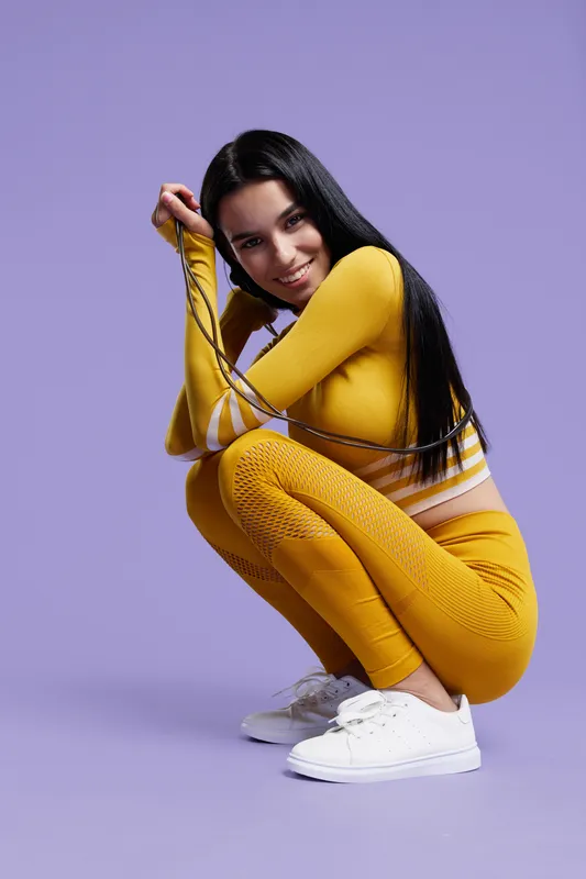 Sport fitness woman in bright yellow activewear crouching with jump rope, smiling confidently against a soft purple studio background, showcasing health and energy.