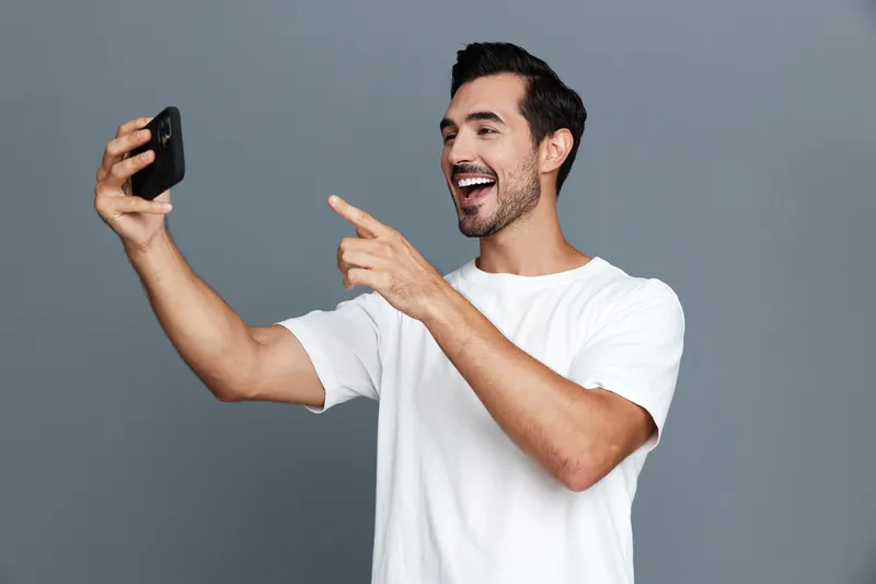 Smiling man wearing a white T-shirt happily looking at his mobile phone while taking a selfie against a plain gray background. Modern lifestyle concept.