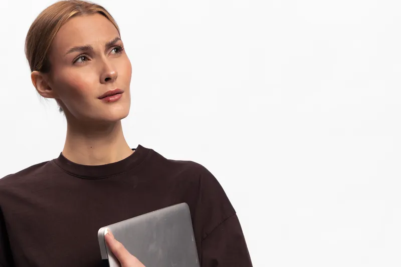 Thoughtful young woman in dark sweater holding tablet looks up against white background. Concept for technology, education, business, and digital communication.