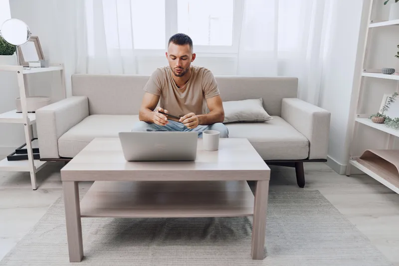 Focused man working on a laptop in a modern living room, surrounded by soft natural light. Casual attire and a cozy atmosphere enhance productivity and creativity.