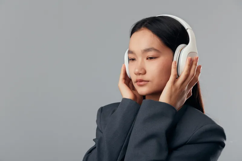 Young woman with closed eyes wearing white headphones and dark jacket listening to music or podcast on gray background. Relaxation, technology, sound and lifestyle concept.