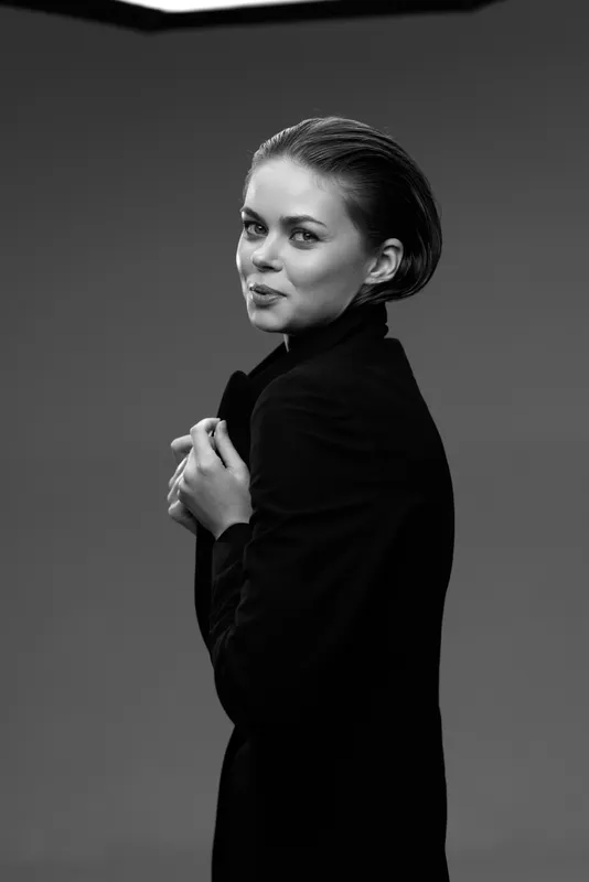 Black and white portrait of a confident woman with short hair wearing a dark coat, showing subtle smile and poised elegance in a studio setting.