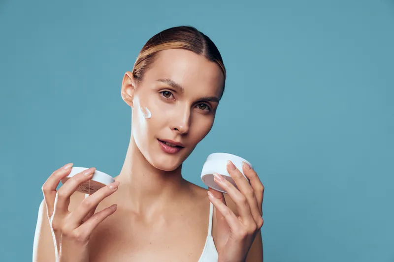 A young woman with clear skin applies white cream on her face holding a jar and finger against a blue background. Concept for skincare, beauty, and daily routine.