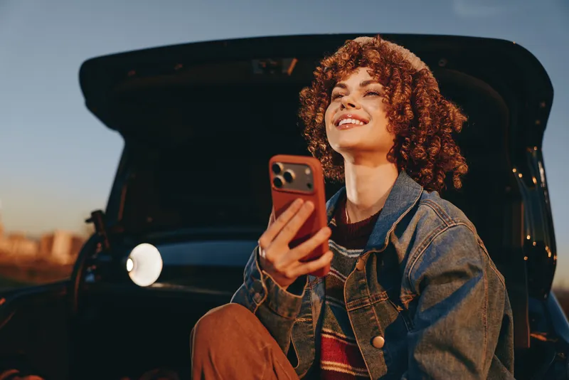 Woman lifestyle smartphone car trunk curly hair smiling, seated in an open car trunk, holding a smartphone in denim jacket, casual travel leisure, evening light, relaxed expression today