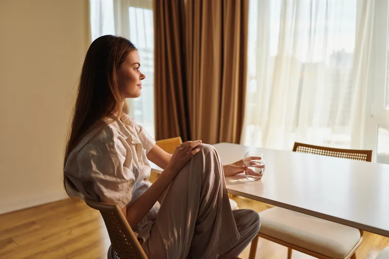 Woman sitting portrait at home by a sunlit table, holding a glass of water and looking thoughtful and relaxed near window, morning calm and cozy interior scene.
