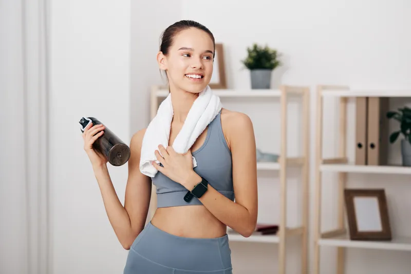Woman in athletic wear with towel around neck, holding a water bottle during workout at home gym. Fitness routine, focus, energy and motivation.