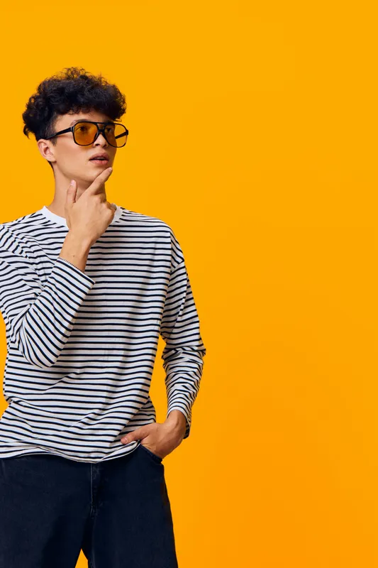 young man wearing yellow sunglasses and striped shirt poses thoughtfully against bright orange background with casual style and curly hair, modern urban fashion vibe