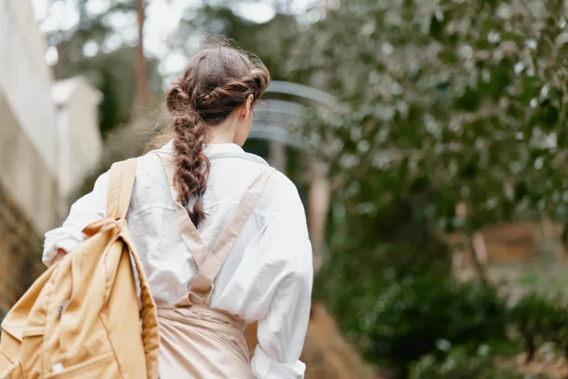 Young woman in casual attire walking outdoors, showcasing a braided hairstyle and a backpack, surrounded by greenery, conveying a sense of adventure and tranquility