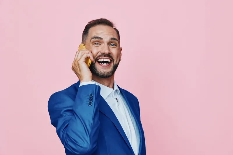 A confident business professional on a pink studio backdrop, wearing a sharp blue suit, talking on a phone with a vivid, joyful expression. The isolated colored background enhances a dynamic, upbeat