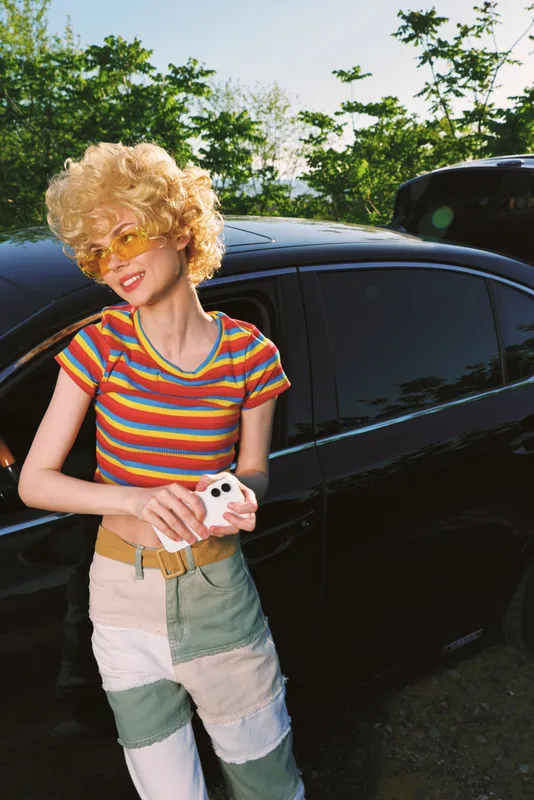 Young woman with curly blond hair stands beside a glossy black car, wearing a striped retro tee and patchwork jeans, smiling softly in a sunlit outdoor moment.