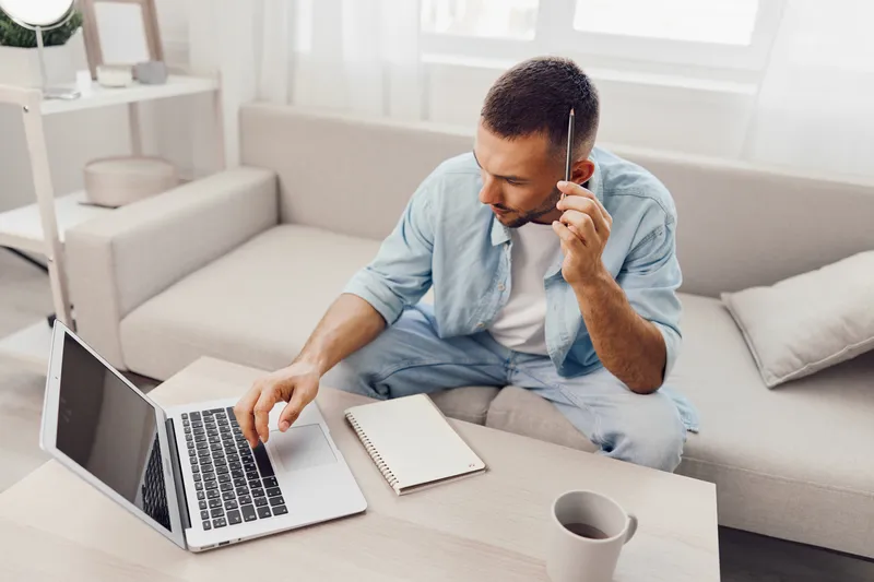 Focused man working on a laptop while taking notes in a cozy, bright living room, showcasing productivity and a modern workspace atmosphere.