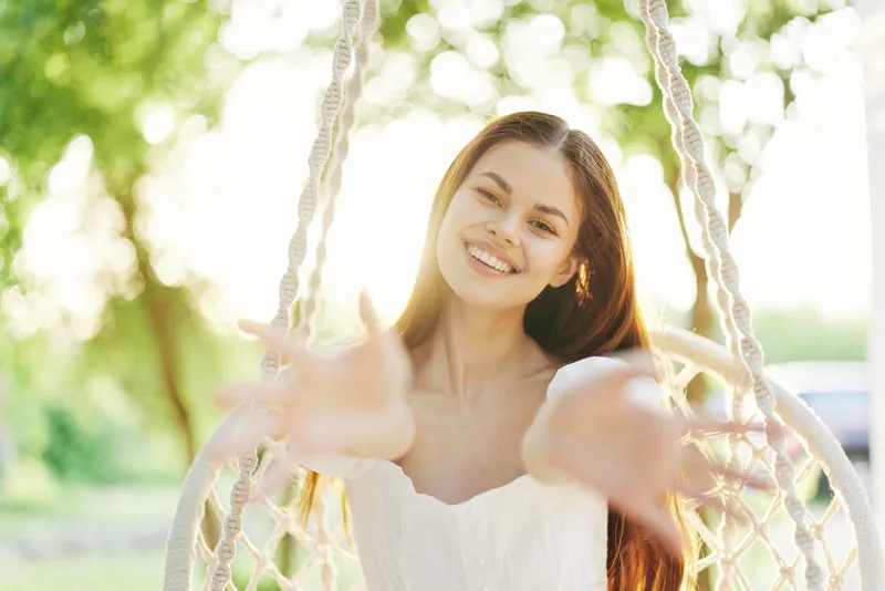 A joyful young woman with long hair in a white dress stretches her arms out while sitting on a swing, surrounded by a sunny green background