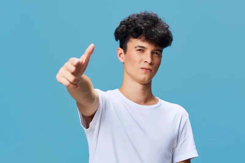 young man pointing with confidence wearing casual white t-shirt against plain blue background expressing assertiveness and determination