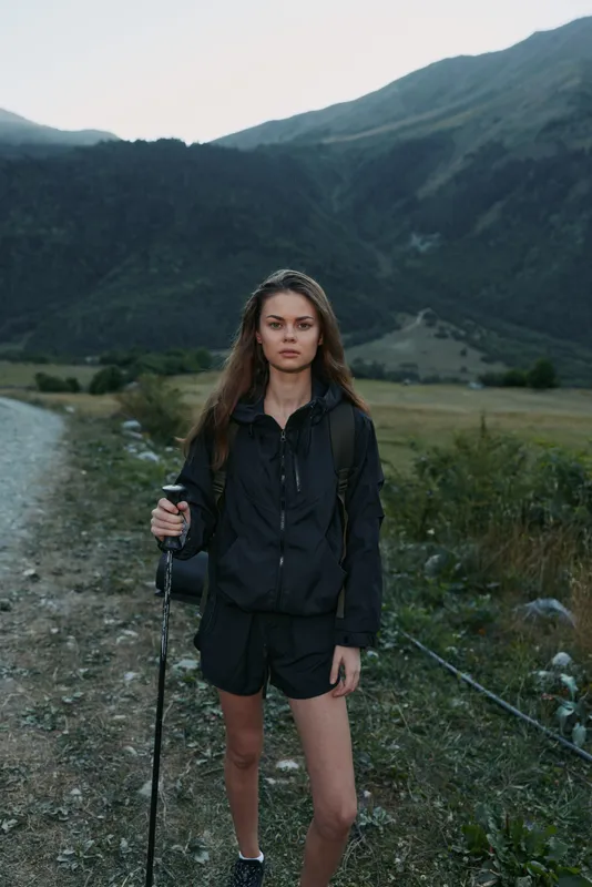 A young female hiker stands in a mountain valley, wearing a dark jacket and shorts, with a trekking pole in hand and a backpack. She gazes ahead with calm, confident focus.