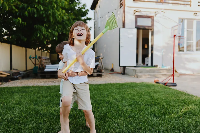 Joyful boys playing outside in a sunny backyard, one holding a rake while the other laughs. They embody carefree childhood moments and playful energy.