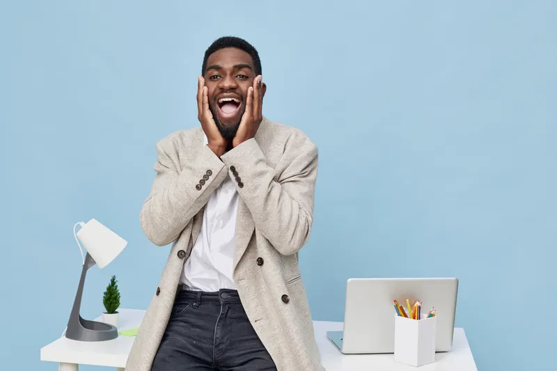 Joyful smiling man with dark skin, short hair, wearing beige blazer and white shirt, standing at modern office desk with laptop, lamp, and plant on light blue background. People lifestyle concept, hap